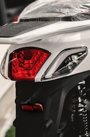 A close-up view of a motorcycle's rear section, highlighting the red tail light and part of the seat. The seat is wrapped in a protective plastic cover, and the suspension spring is visible.