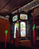 An ornate Chinese lantern with intricate black carvings and illuminated panels displaying traditional paintings of trees, houses, and a rising sun. The lantern is adorned with red tassels and green decorative knots, creating a festive and cultural ambiance.