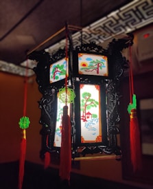 An ornate Chinese lantern with intricate black carvings and illuminated panels displaying traditional paintings of trees, houses, and a rising sun. The lantern is adorned with red tassels and green decorative knots, creating a festive and cultural ambiance.