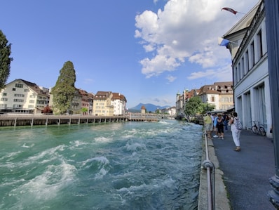 A riverside scene with a fast-flowing, turquoise river bordered by historic buildings. The sky is clear with a few scattered, fluffy clouds. People are standing along the promenade, taking in the view. Flags are fluttering atop the buildings on the right, and a distant bridge and mountain range can be seen.
