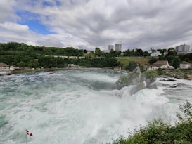 A powerful waterfall cascades over rocky terrain, surrounded by lush greenery and trees. In the background, there are several buildings and infrastructure, including a bridge with arches and a few modern high-rise buildings under construction. A Swiss flag is visible in the foreground.