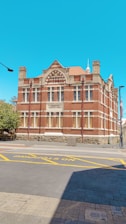 A historic red brick building with intricate architectural details like arched windows and stone accents. The building is labeled 'Fremantle Technical School'. It stands on a paved street with markings for no stopping zones, against a bright blue sky.