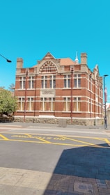 A historic red brick building with intricate architectural details like arched windows and stone accents. The building is labeled 'Fremantle Technical School'. It stands on a paved street with markings for no stopping zones, against a bright blue sky.