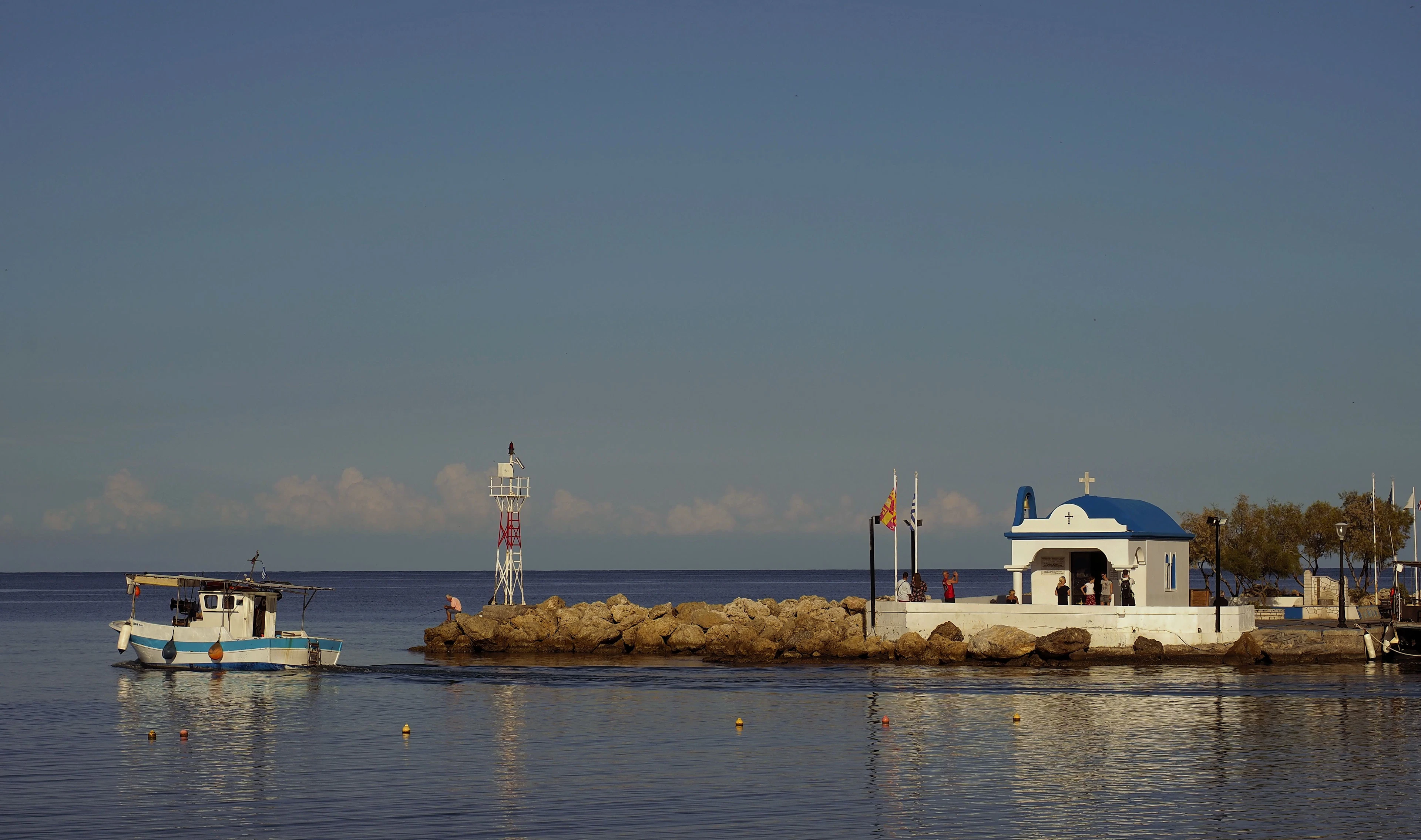 A small white chapel with a blue dome on a stone breakwater beside a calm harbor, with a fishing boat moored nearby.