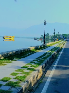 A tranquil lakeside scene with a paved walkway alongside a calm body of water. A yellow houseboat is reflected in the water, and the distant shoreline is lined with trees. Several lampposts line the walkway next to a road with a white road marking. The scene has a backdrop of hazy mountains under a clear sky.