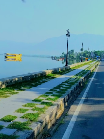 A tranquil lakeside scene with a paved walkway alongside a calm body of water. A yellow houseboat is reflected in the water, and the distant shoreline is lined with trees. Several lampposts line the walkway next to a road with a white road marking. The scene has a backdrop of hazy mountains under a clear sky.