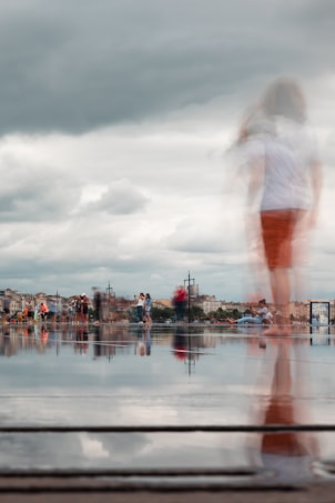 People are gathered around a reflective surface, likely a shallow pool or wet pavement, with an urban landscape in the background. The scene appears to be taken on a cloudy day, and there is a blurred effect of someone walking, suggesting motion.