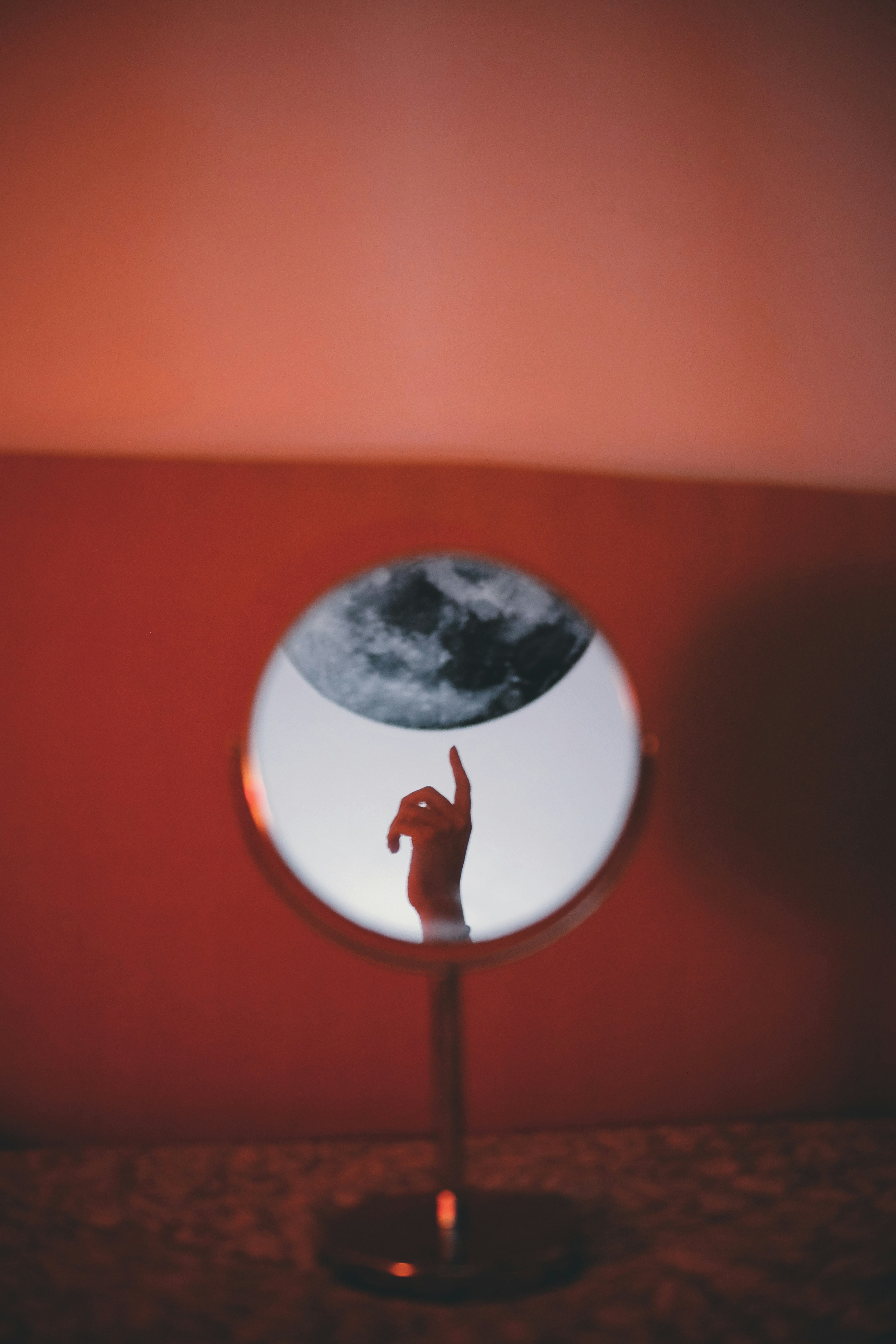 Circular mirror on a stand reflects a hand with an extended finger and a moon-like shape against a red wall.