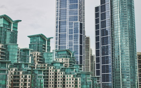 A cluster of modern skyscrapers with a contemporary architectural style featuring glass facades and geometric shapes. The buildings have a bluish-green tint, creating a sleek and urban atmosphere.