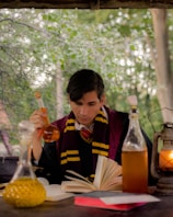 A person in a black robe with maroon and yellow stripes is intently reading a book at a wooden table. Surrounding the person are multiple glass bottles filled with colorful liquids. A lantern with a burning flame adds a warm glow to the scene. Behind is a backdrop of lush green trees and leaves.
