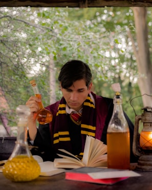 A person in a black robe with maroon and yellow stripes is intently reading a book at a wooden table. Surrounding the person are multiple glass bottles filled with colorful liquids. A lantern with a burning flame adds a warm glow to the scene. Behind is a backdrop of lush green trees and leaves.