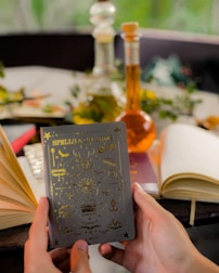 A pair of hands holds a book titled 'Spells & Charms,' adorned with golden symbols on the cover. Surrounding the book are several open and closed notebooks on a table. In the background, there are two glass bottles containing colored liquids, possibly potion ingredients. Greenery and blurred natural surroundings add a serene and mystical ambiance.