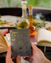 A pair of hands holds a book titled 'Spells & Charms,' adorned with golden symbols on the cover. Surrounding the book are several open and closed notebooks on a table. In the background, there are two glass bottles containing colored liquids, possibly potion ingredients. Greenery and blurred natural surroundings add a serene and mystical ambiance.