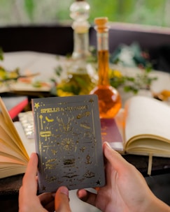A pair of hands holds a book titled 'Spells & Charms,' adorned with golden symbols on the cover. Surrounding the book are several open and closed notebooks on a table. In the background, there are two glass bottles containing colored liquids, possibly potion ingredients. Greenery and blurred natural surroundings add a serene and mystical ambiance.
