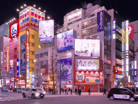 A vibrant urban street scene featuring colorful illuminated billboards and advertisements on multi-story buildings. The street is lively with people walking and cars driving by, set against a backdrop of a bustling cityscape at night.