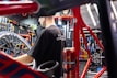 A person is working on a bicycle in a well-organized workshop. The area is filled with various tools neatly arranged on a pegboard. A red bicycle frame and wheel are prominently visible, and there is a can on the workbench.