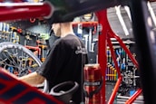 A person is working on a bicycle in a well-organized workshop. The area is filled with various tools neatly arranged on a pegboard. A red bicycle frame and wheel are prominently visible, and there is a can on the workbench.