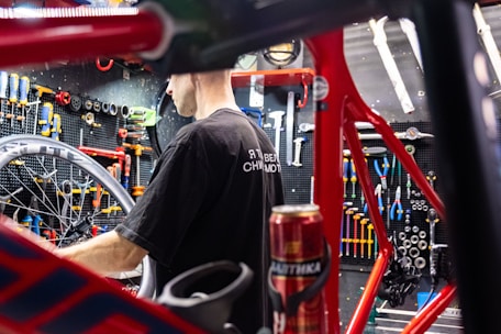 A person is working on a bicycle in a well-organized workshop. The area is filled with various tools neatly arranged on a pegboard. A red bicycle frame and wheel are prominently visible, and there is a can on the workbench.