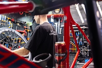 A person is working on a bicycle in a well-organized workshop. The area is filled with various tools neatly arranged on a pegboard. A red bicycle frame and wheel are prominently visible, and there is a can on the workbench.