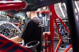 A person is working on a bicycle in a well-organized workshop. The area is filled with various tools neatly arranged on a pegboard. A red bicycle frame and wheel are prominently visible, and there is a can on the workbench.