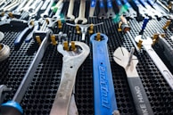 A collection of various wrenches and hand tools neatly arranged on a black perforated surface. The tools are of different sizes and colors, including a prominently visible large blue wrench in the center. The arrangement suggests an organized workspace or tool display.