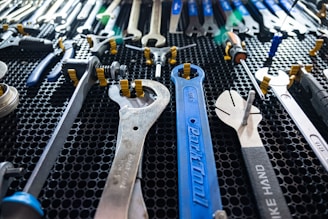 A collection of various wrenches and hand tools neatly arranged on a black perforated surface. The tools are of different sizes and colors, including a prominently visible large blue wrench in the center. The arrangement suggests an organized workspace or tool display.