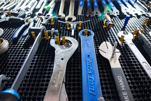 A collection of various wrenches and hand tools neatly arranged on a black perforated surface. The tools are of different sizes and colors, including a prominently visible large blue wrench in the center. The arrangement suggests an organized workspace or tool display.