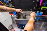 A person with a star tattoo on their arm operates an ultrasonic cleaner. They are cleaning a bicycle cassette in a workshop with various tools hanging on a black pegboard. A large bottle of cleaning solution is on the counter nearby.