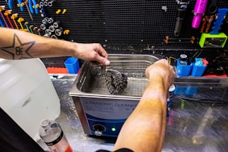 A person with a star tattoo on their arm operates an ultrasonic cleaner. They are cleaning a bicycle cassette in a workshop with various tools hanging on a black pegboard. A large bottle of cleaning solution is on the counter nearby.