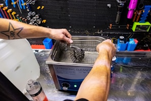A person with a star tattoo on their arm operates an ultrasonic cleaner. They are cleaning a bicycle cassette in a workshop with various tools hanging on a black pegboard. A large bottle of cleaning solution is on the counter nearby.