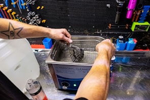 A person with a star tattoo on their arm operates an ultrasonic cleaner. They are cleaning a bicycle cassette in a workshop with various tools hanging on a black pegboard. A large bottle of cleaning solution is on the counter nearby.