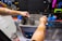 A person with a star tattoo on their arm operates an ultrasonic cleaner. They are cleaning a bicycle cassette in a workshop with various tools hanging on a black pegboard. A large bottle of cleaning solution is on the counter nearby.