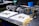 A workbench features various mechanical parts, including metal components and bolts, arranged on white cloths. The setting appears to be a workshop or garage with tools and equipment in the background, such as a machine in green and a blue container. The metal objects seem to be automotive or machinery-related, each with intricate design details.