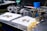 A workbench features various mechanical parts, including metal components and bolts, arranged on white cloths. The setting appears to be a workshop or garage with tools and equipment in the background, such as a machine in green and a blue container. The metal objects seem to be automotive or machinery-related, each with intricate design details.