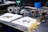 A workbench features various mechanical parts, including metal components and bolts, arranged on white cloths. The setting appears to be a workshop or garage with tools and equipment in the background, such as a machine in green and a blue container. The metal objects seem to be automotive or machinery-related, each with intricate design details.