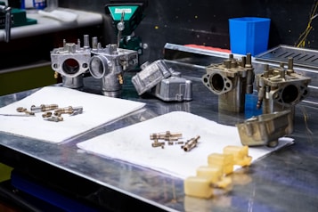 A workbench features various mechanical parts, including metal components and bolts, arranged on white cloths. The setting appears to be a workshop or garage with tools and equipment in the background, such as a machine in green and a blue container. The metal objects seem to be automotive or machinery-related, each with intricate design details.
