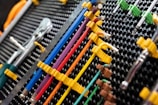 A collection of neatly organized hand tools, including wrenches and screwdrivers, is arranged on a perforated panel. The tools have colorful handles in shades of yellow, blue, red, and purple, which are secured in position by yellow fasteners. The background pattern is composed of a series of black circular holes, adding a textured look to the arrangement.