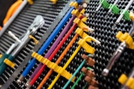 A collection of neatly organized hand tools, including wrenches and screwdrivers, is arranged on a perforated panel. The tools have colorful handles in shades of yellow, blue, red, and purple, which are secured in position by yellow fasteners. The background pattern is composed of a series of black circular holes, adding a textured look to the arrangement.