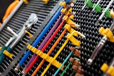 A collection of neatly organized hand tools, including wrenches and screwdrivers, is arranged on a perforated panel. The tools have colorful handles in shades of yellow, blue, red, and purple, which are secured in position by yellow fasteners. The background pattern is composed of a series of black circular holes, adding a textured look to the arrangement.