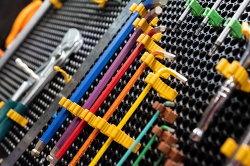 A collection of neatly organized hand tools, including wrenches and screwdrivers, is arranged on a perforated panel. The tools have colorful handles in shades of yellow, blue, red, and purple, which are secured in position by yellow fasteners. The background pattern is composed of a series of black circular holes, adding a textured look to the arrangement.