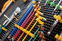 A collection of neatly organized hand tools, including wrenches and screwdrivers, is arranged on a perforated panel. The tools have colorful handles in shades of yellow, blue, red, and purple, which are secured in position by yellow fasteners. The background pattern is composed of a series of black circular holes, adding a textured look to the arrangement.