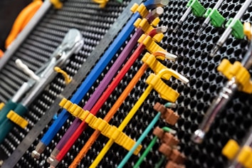 A collection of neatly organized hand tools, including wrenches and screwdrivers, is arranged on a perforated panel. The tools have colorful handles in shades of yellow, blue, red, and purple, which are secured in position by yellow fasteners. The background pattern is composed of a series of black circular holes, adding a textured look to the arrangement.