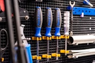 A wall mounted tool organizer holds a set of screwdrivers with blue and black handles, secured with yellow clamps. Additional tools, including a wrench and metal bars, are organized neatly against a perforated black metal background.