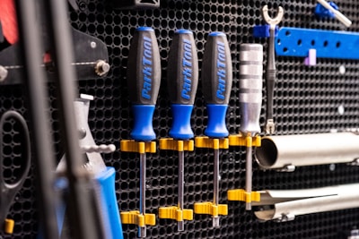 A wall mounted tool organizer holds a set of screwdrivers with blue and black handles, secured with yellow clamps. Additional tools, including a wrench and metal bars, are organized neatly against a perforated black metal background.