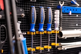 A wall mounted tool organizer holds a set of screwdrivers with blue and black handles, secured with yellow clamps. Additional tools, including a wrench and metal bars, are organized neatly against a perforated black metal background.