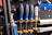 A wall mounted tool organizer holds a set of screwdrivers with blue and black handles, secured with yellow clamps. Additional tools, including a wrench and metal bars, are organized neatly against a perforated black metal background.