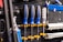 A wall mounted tool organizer holds a set of screwdrivers with blue and black handles, secured with yellow clamps. Additional tools, including a wrench and metal bars, are organized neatly against a perforated black metal background.