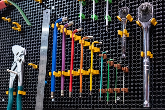 A neatly organized set of tools displayed on a wall-mounted pegboard. The tools include a set of colored hex keys, pliers, a metal ruler, various ratchets, and sockets. The tools are held in place by yellow clips attached to the perforated board. The predominant background is a black, hexagonal grid pattern, with additional green clips holding some of the tools.