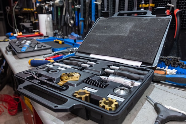 A black toolbox is open on a cluttered workbench, containing various metallic tools and components. Other tools, including screwdrivers and pliers, are scattered across the surface. The workspace appears to be in a workshop or garage setting, with additional equipment hanging on the wall in the background.