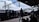 The image depicts a busy train station platform with several people waiting. A long freight train is passing by on the tracks, bearing industrial markings and logos. Overhead, an elevated footbridge connects different parts of the station. The platform's roofing offers partial shade to passengers, blending with the industrial ambiance of the setting.